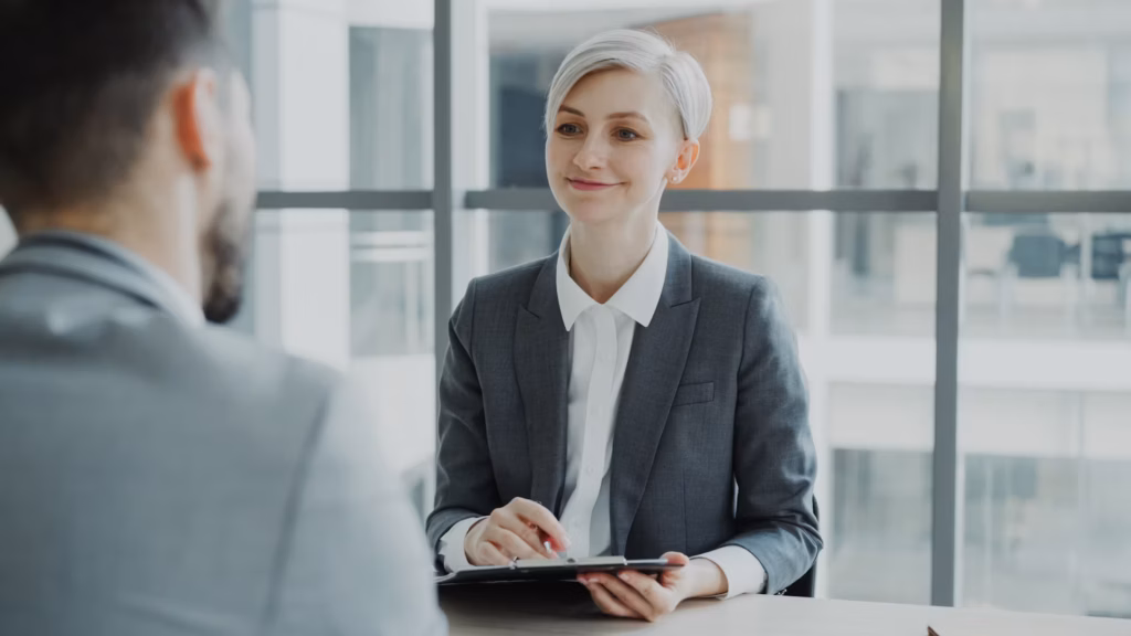 Professional woman smiling and interviewing a candidate in an office setting, holding a clipboard during a job interview.