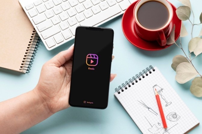 Hand holding a smartphone displaying the Instagram Reels logo on a desk with a coffee cup, keyboard, notebook, and pen.