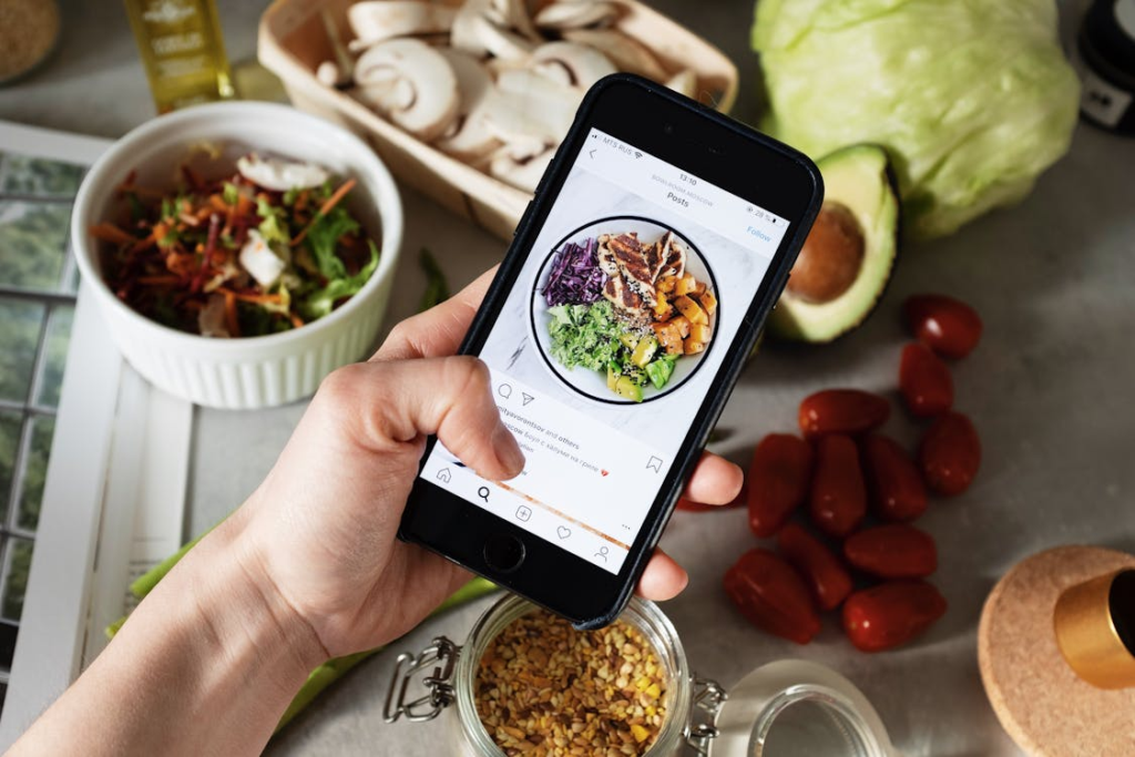 Hand holding a smartphone displaying a healthy meal Instagram post, surrounded by fresh ingredients like avocado, mushrooms, lettuce, and cherry tomatoes on a kitchen counter.