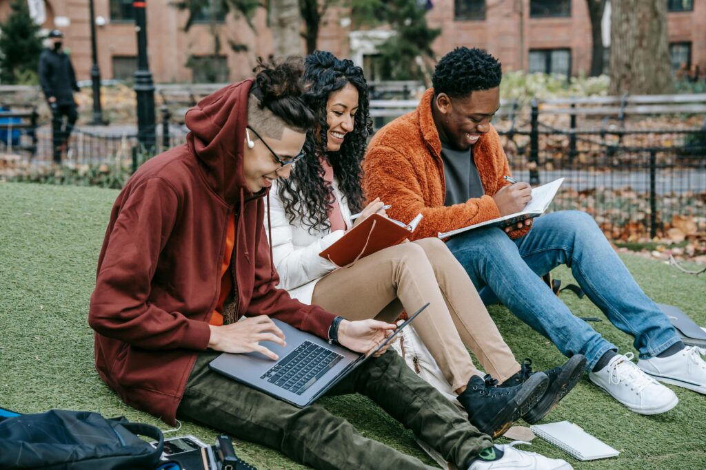 Three college students sitting on grass in a park studying together, using a laptop and notebooks, smiling while working on assignments, representing collaborative learning, university life, group study, and academic teamwork outdoors.
