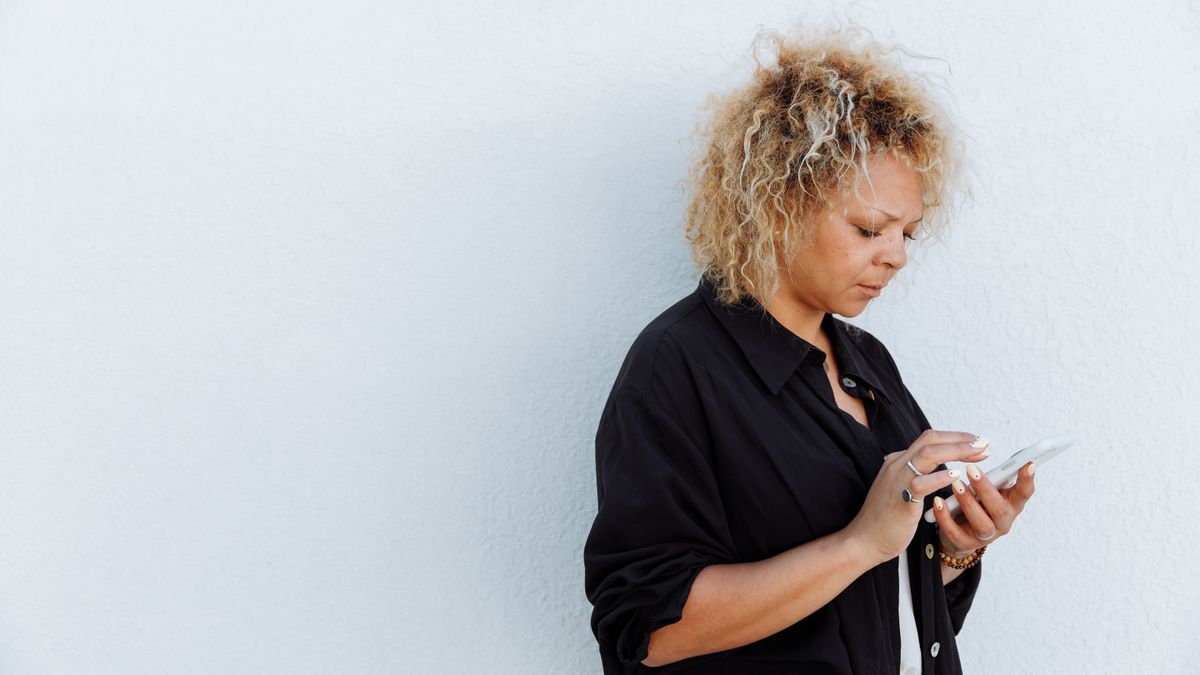 A woman with curly hair uses a smartphone against a white wall outdoors.