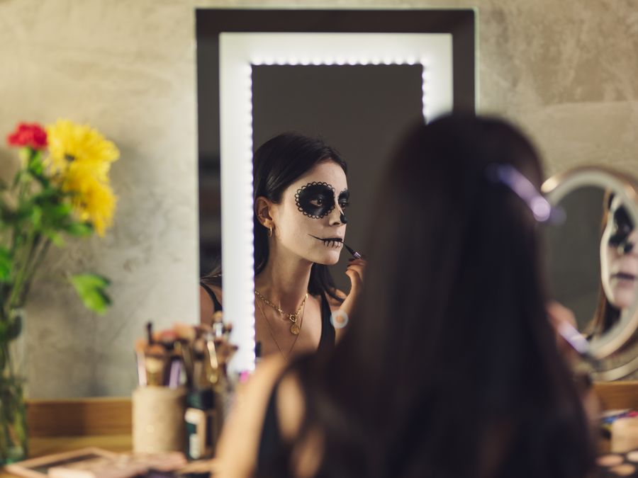Woman applying Catrina makeup for Day of the Dead in front of a mirror adorned with flowers.