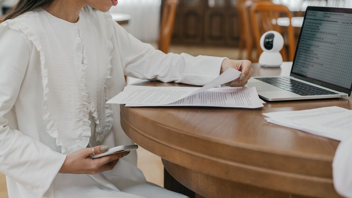 Woman in white dress studying at a wooden table with a laptop and papers indoors.