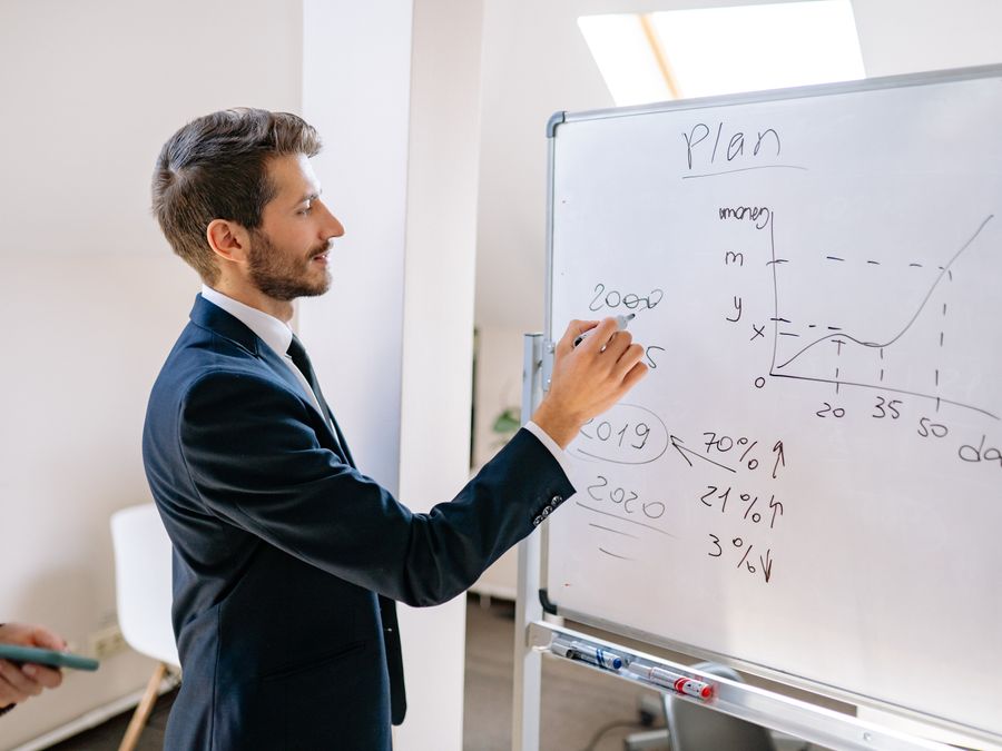 A businessman in a suit writes financial data on a whiteboard during an office planning session.
