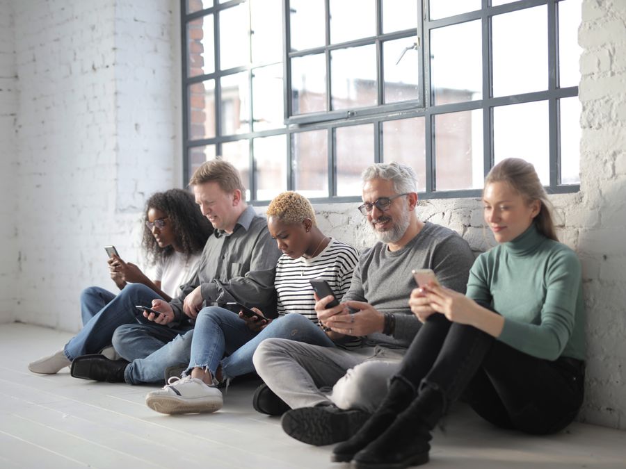 Smiling coworkers of different ages in casual outfit browsing internet on cellphones while sitting with crossed legs near brick wall and fenced window during daytime
