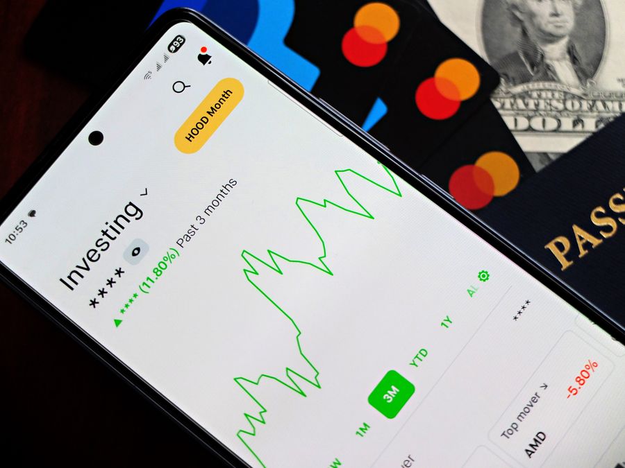 A smartphone showing an investment app with green growth indicators, surrounded by credit cards, US dollars, and a passport.