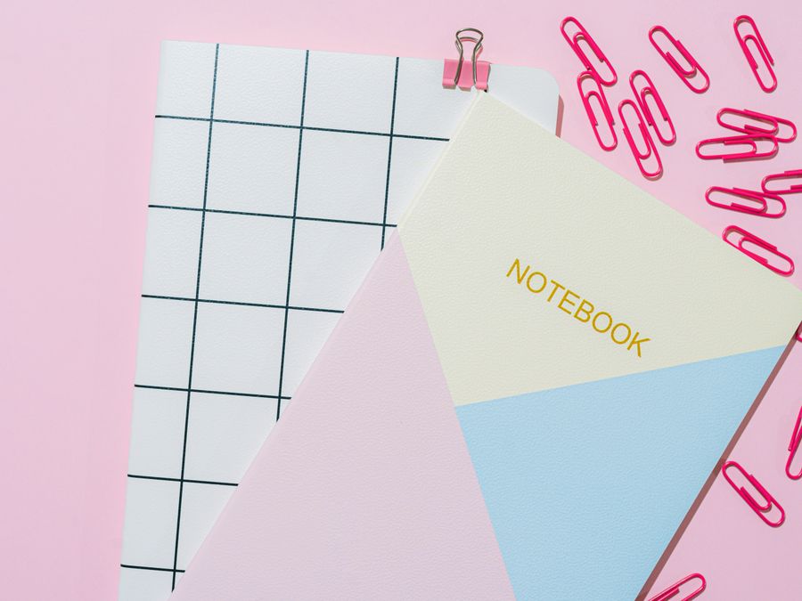 Flat lay of notebooks, paper clips, and binder clip on a pink background.
