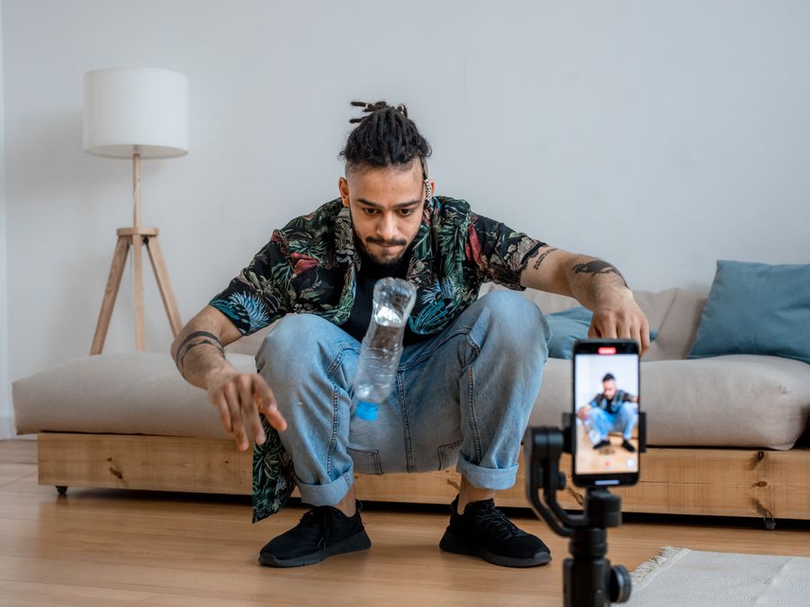 A young man focuses on a bottle flip challenge while filming content at home, capturing it on a smartphone.