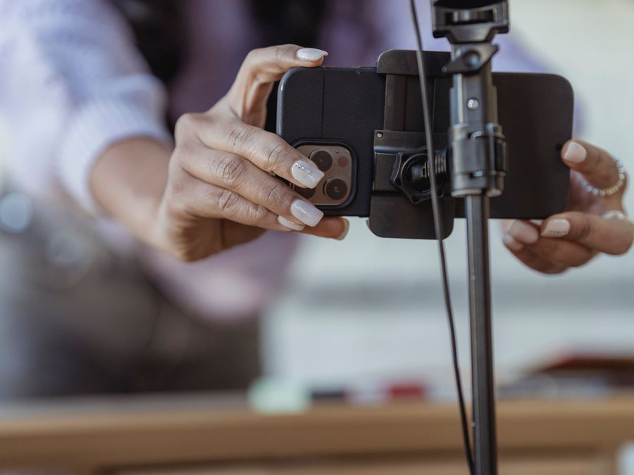 Crop unrecognizable female setting mobile phone on tripod while recording vlog on blurred background