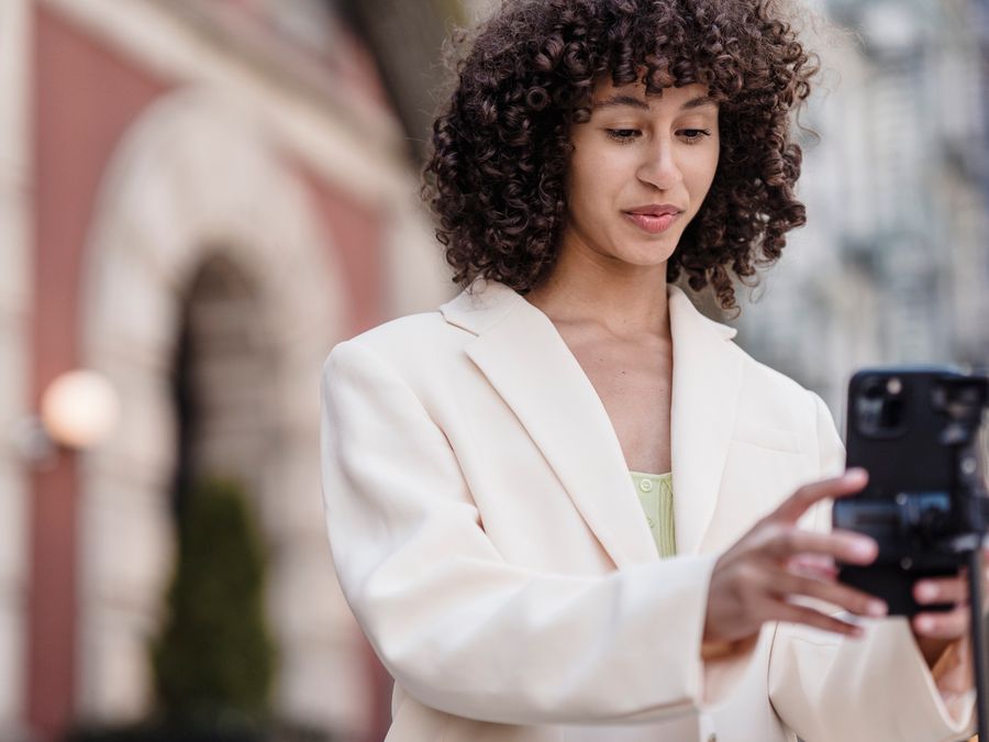 Young ethnic female vlogger with curly hair in jacket preparing cellphone for video record in town