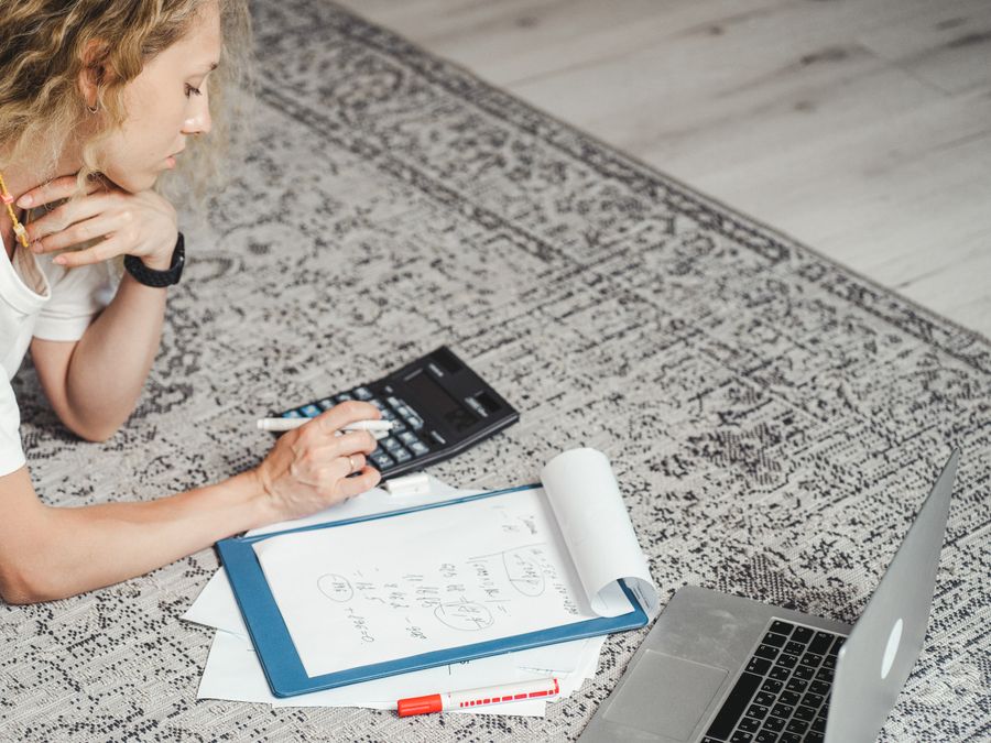 A woman calculates finances at home using a laptop, calculator, and documents on a rug.