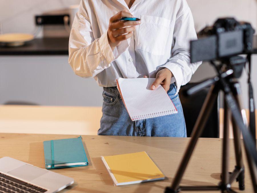 Vlogger recording content with a camera, holding notes and using a laptop in a modern setting.