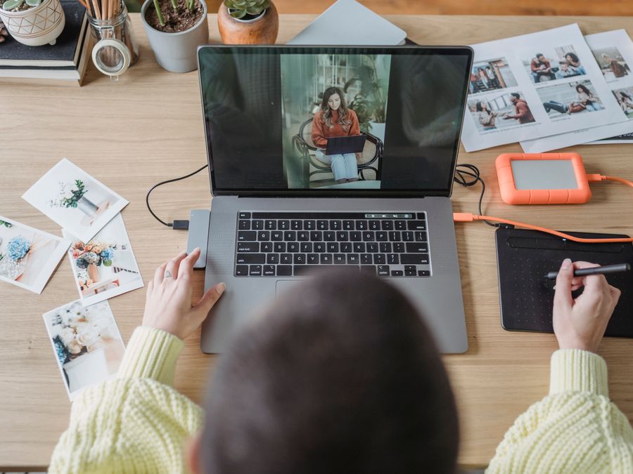 From above of crop faceless female photographer drawing on graphic tablet while editing photos on laptop at table with printed photos