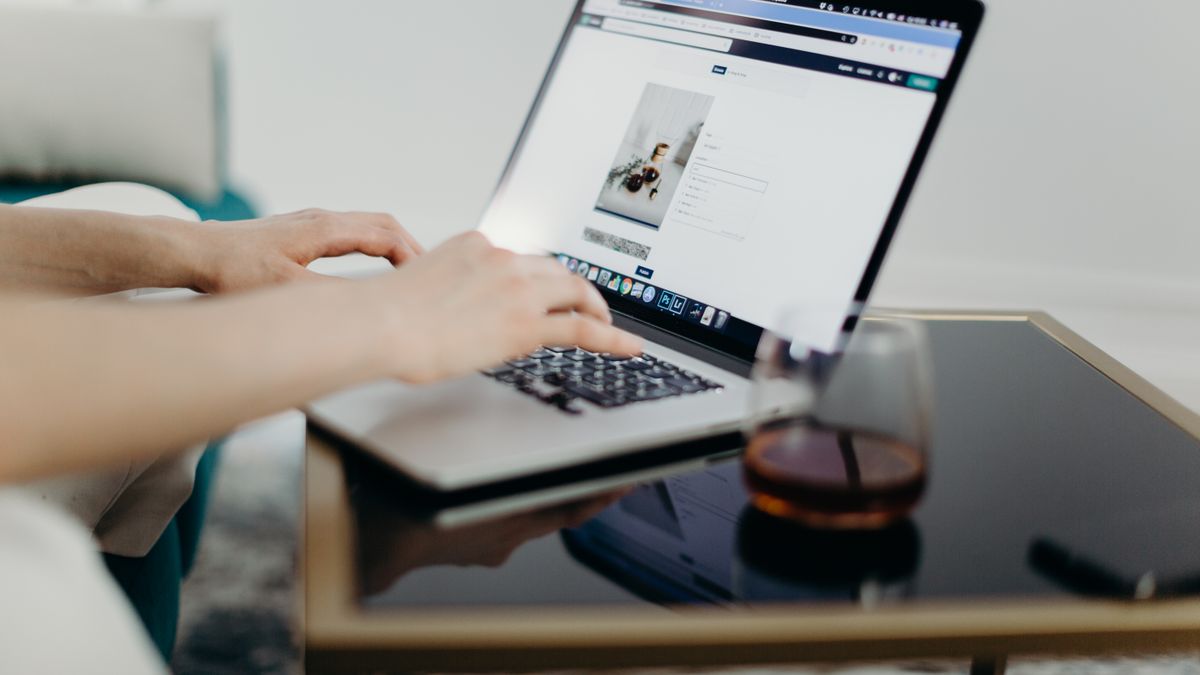 A person typing on a laptop in a stylish home office with a glass of drink nearby.