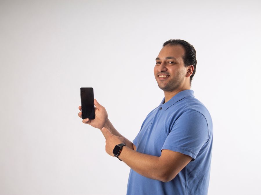 Smiling man in blue shirt holding and pointing to a black smartphone against a white background.