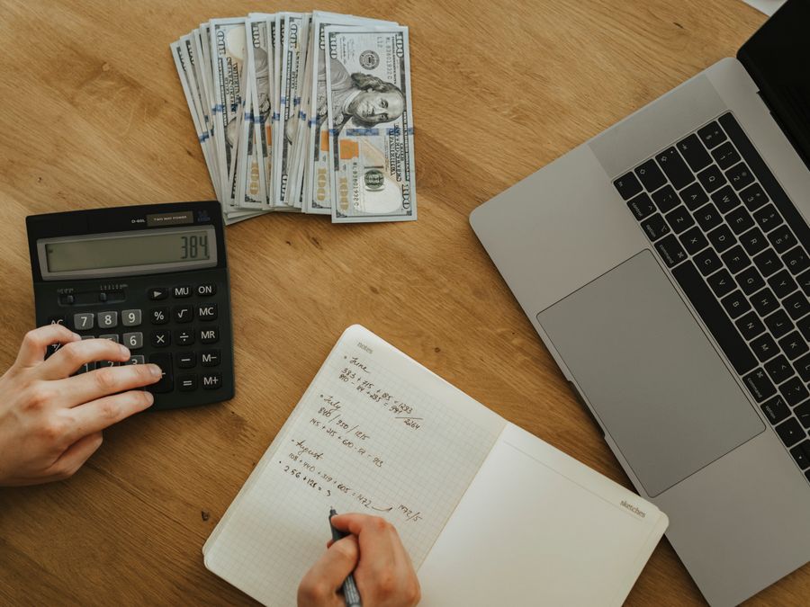 Hands using calculator and notebook, planning finances, with cash and laptop on wooden table.