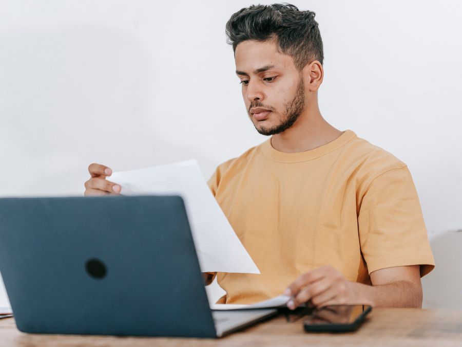 Young bearded Hispanic male university student reading information on paper while studying online via laptop