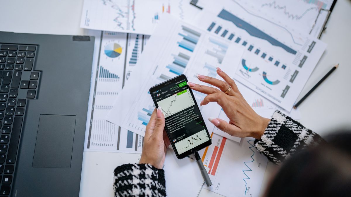 Businesswoman analyzing financial charts on smartphone with laptop and documents.