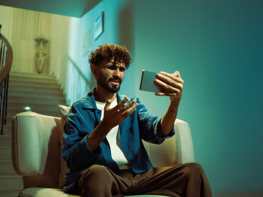 A man sitting on a couch indoors, looking confused while using a smartphone.