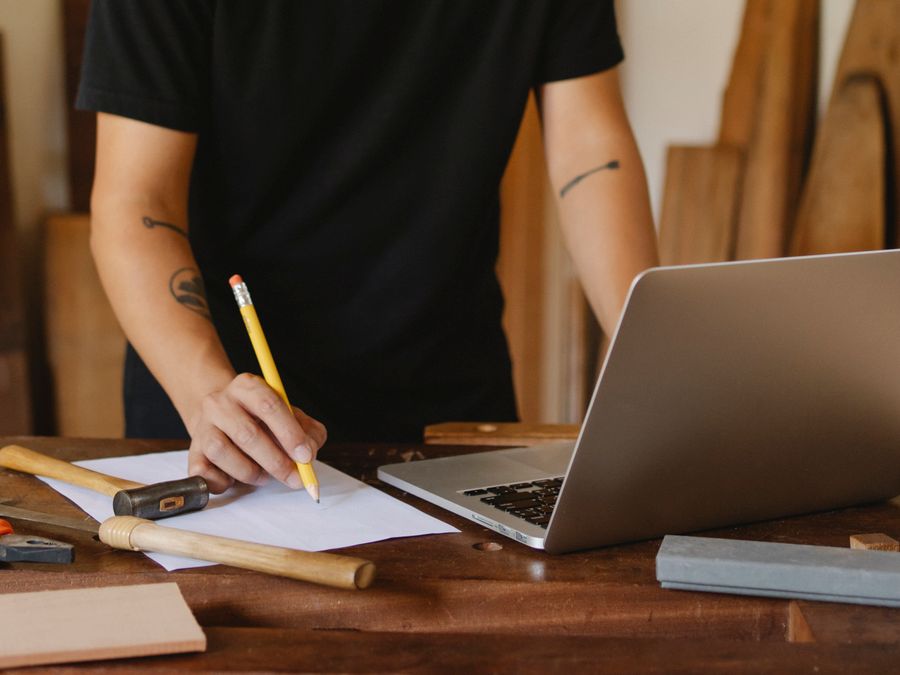 Crop unrecognizable tattooed male master standing at table with various tools and drawing on paper while using laptop during work in carpentry studio