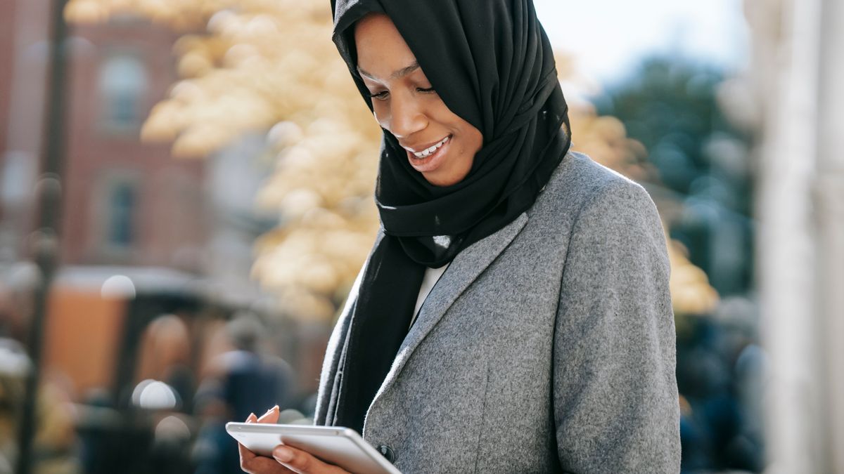 Content young African American Muslim businesswoman in stylish suit and traditional hijab smiling and reading notes on tablet while standing on city street