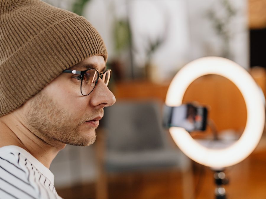 A man wearing eyeglasses and a knitted cap records a video at home using a smartphone and ring light.