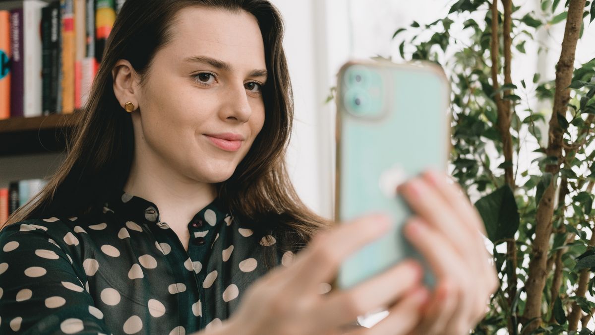 Self assured young female with long dark hair in elegant blouse smiling while taking selfie standing near bookshelf at home