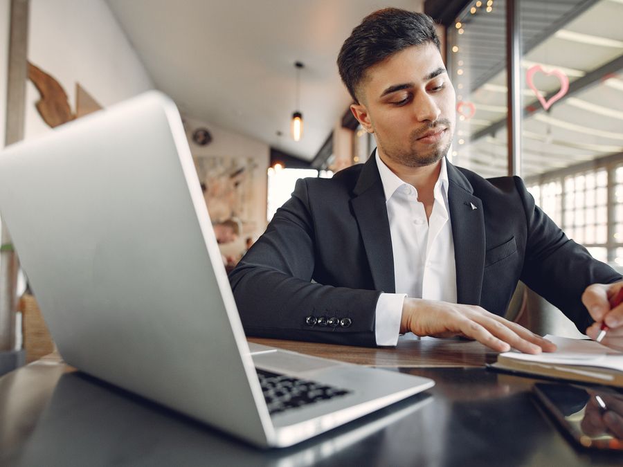 From below of concentrated ethnic male in formal wear sitting in creative workplace and taking notes while using laptop and working on business project