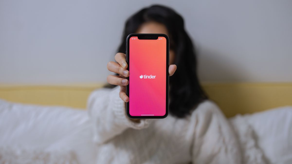 A woman in casual attire displays the Tinder app on her smartphone while sitting indoors.
