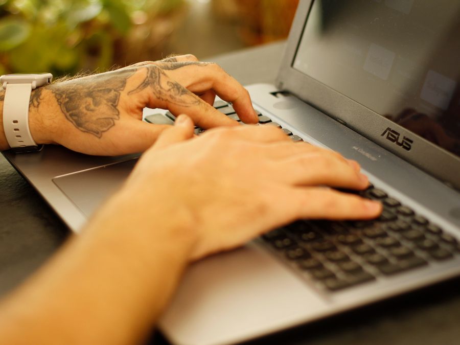 Tattooed hands typing on a laptop keyboard, focused and working.
