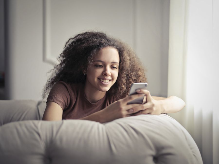 A smiling young woman with curly hair, comfortably texting on her smartphone indoors.