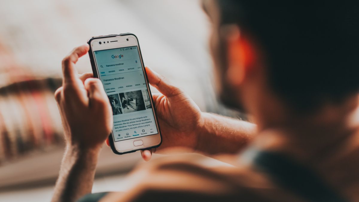 Back view of crop anonymous young male reading news on mobile phone while sitting in bedroom