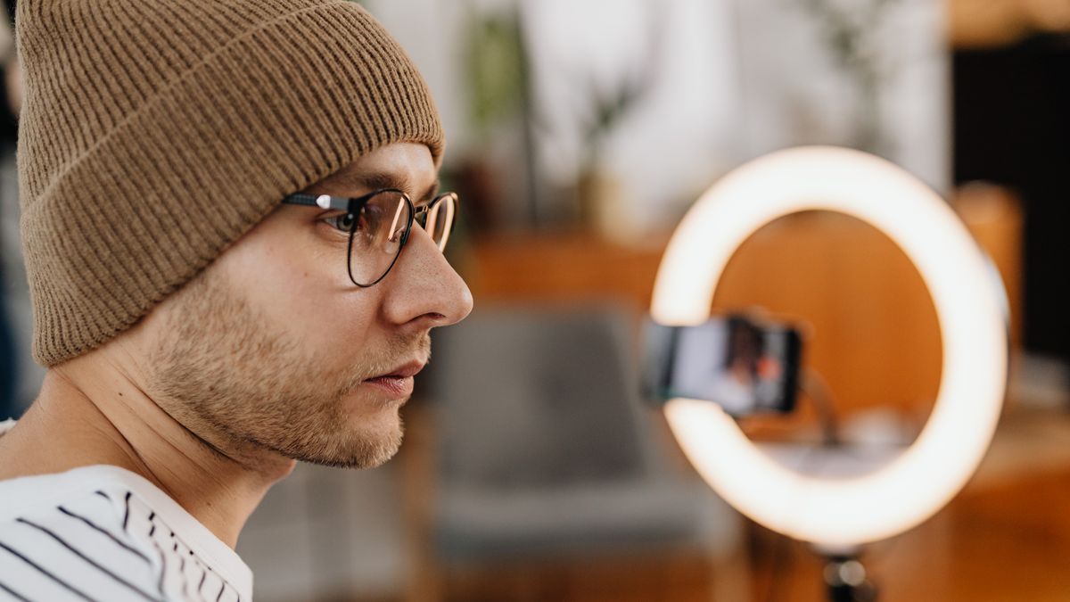 A man wearing eyeglasses and a knitted cap records a video at home using a smartphone and ring light.
