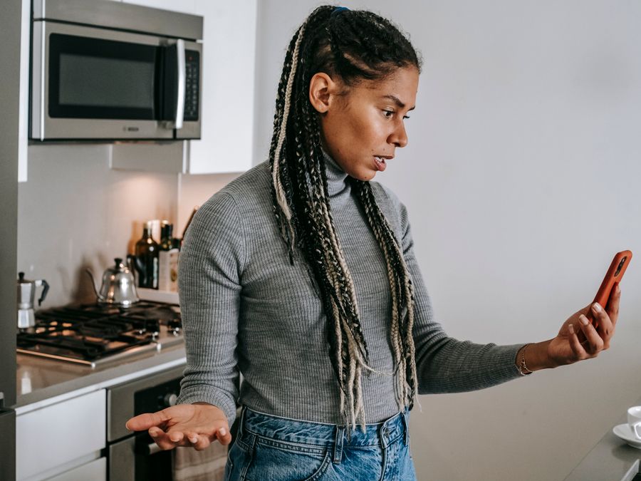 Serious young black lady with Afro braids in casual clothes gesticulating while having unpleasant conversation via video chat on smartphone in modern kitchen