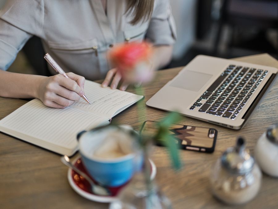 Woman writing in a notebook with a laptop and coffee cup on a desk. Ideal for workspace inspiration.