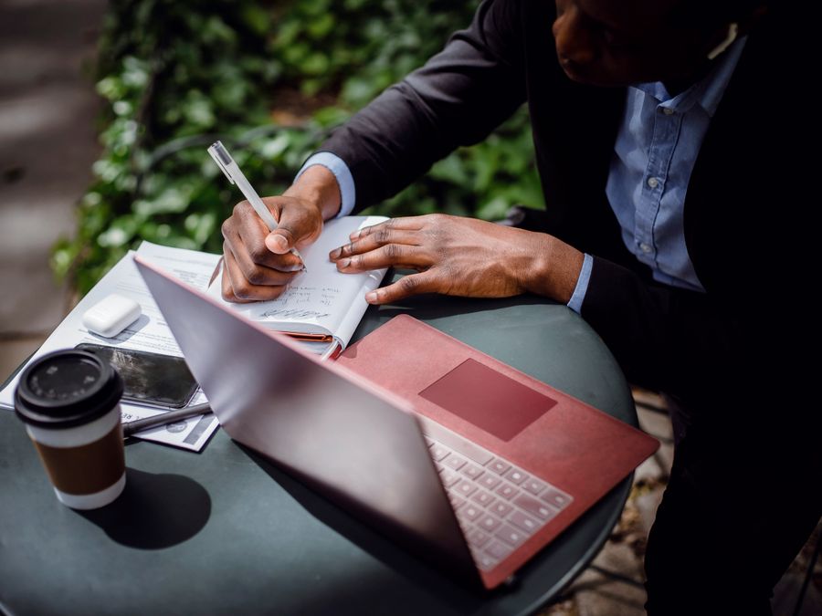 From above of crop black male journalist in formal wear writing article while sitting at table with red laptop and cup of takeaway coffee