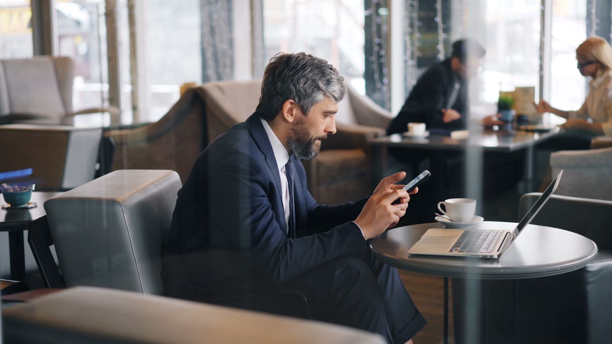 A businessman in a suit using his smartphone and laptop in a modern café setting.