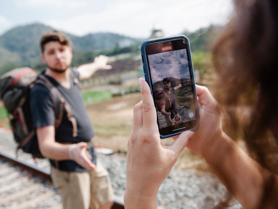 Crop unrecognizable female taking video on smartphone of hiker in casual clothes with backpack standing on railroad and pointing away in countryside in sunny day