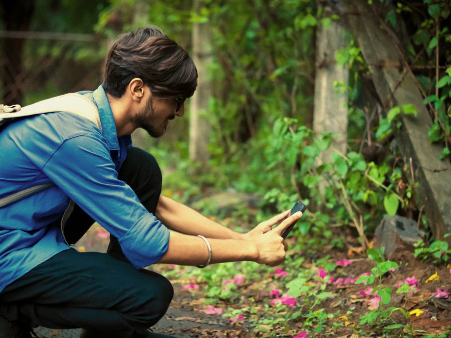 A young man squats outdoors, photographing flowers with a smartphone.