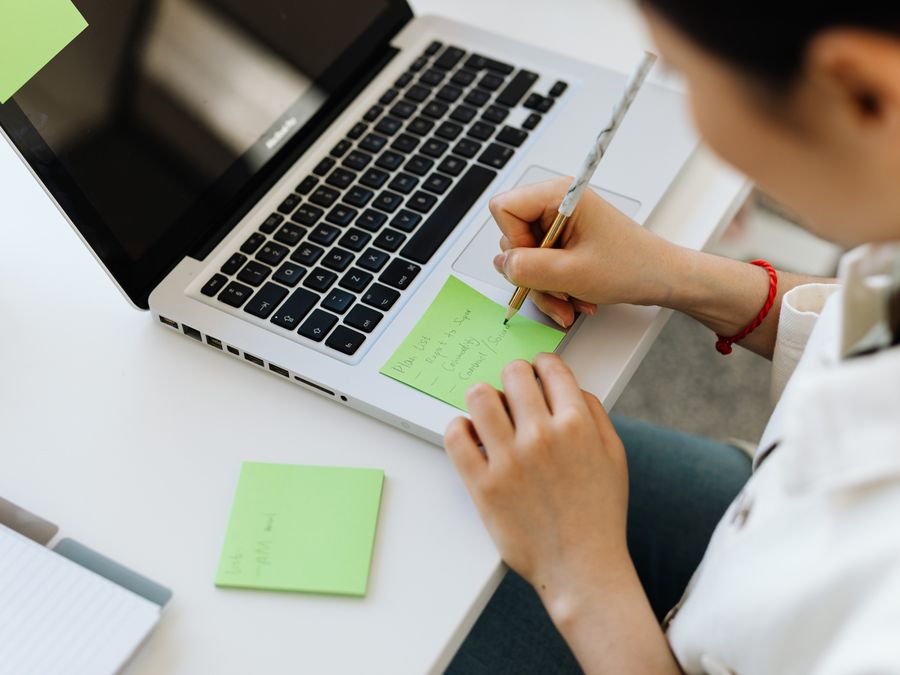 Person writing a to-do list on sticky note next to laptop for organization.