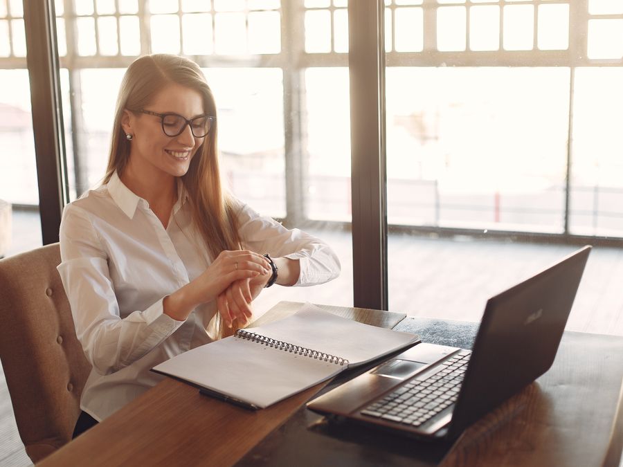 Positive young female entrepreneur in formal clothes and eyeglasses sitting at wooden table with laptop and copybook and checking time on wristwatch while working in light creative workspace