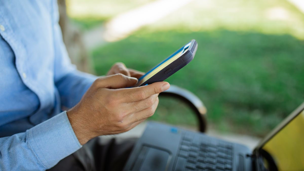 A person uses a smartphone while seated outdoors with a laptop, showcasing remote work lifestyle.