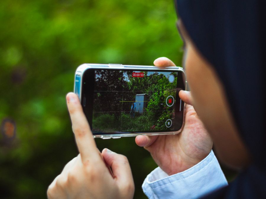 Close-up of person using smartphone to film a green outdoor scene in daylight.