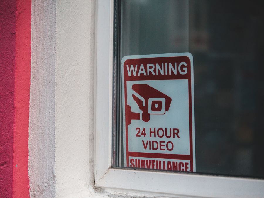 Red warning sign of video surveillance placed behind window of building in daytime