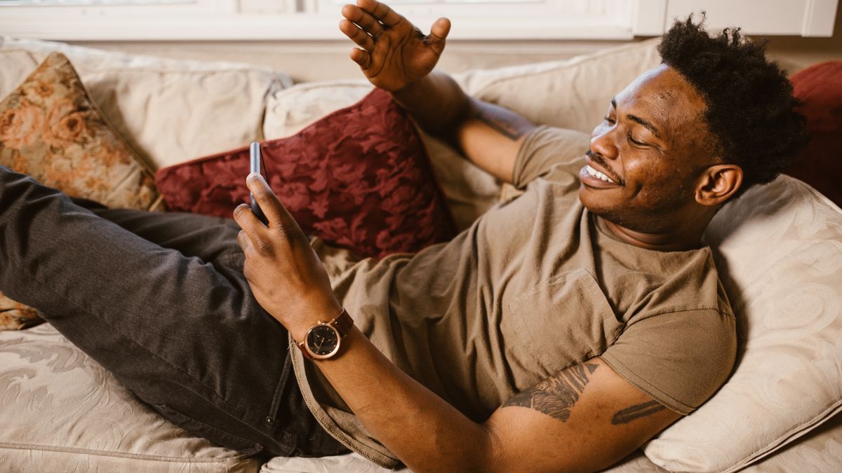 African American man lying on couch, smiling and using smartphone for communication.