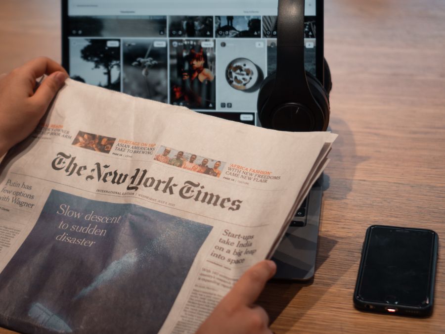 Close-up of hands holding a newspaper by a laptop and smartphone on a table.