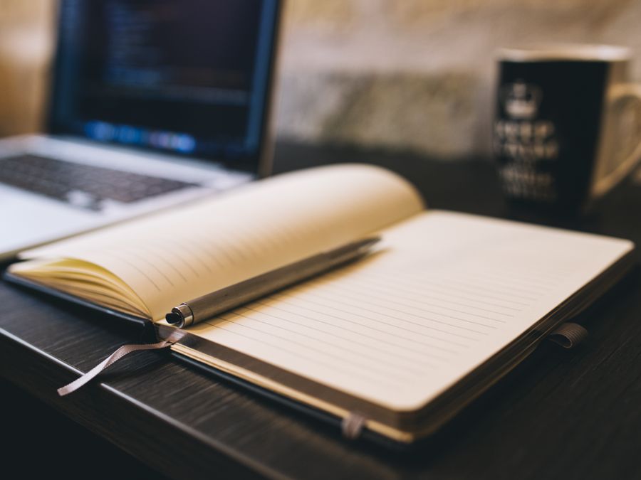Open notebook with a pen on a wooden desk beside a laptop, ideal for blogging and work-related themes.